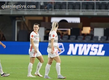 Soccer players in team uniforms stand on a grass field with a large digital advertisement screen displaying the acronym 'NWSL' in the background. The setting appears to be a stadium with rows of empty seats visible above the screen.