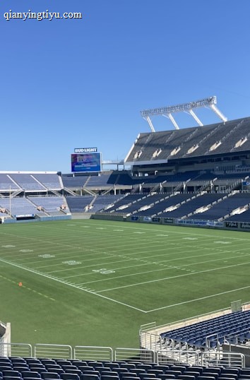A large, empty sports stadium with blue seats surrounding a well-maintained green grass field. Bright sunlight creates clear shadows, and there are two goal posts on opposite ends of the field. A big electronic scoreboard displays some graphics, and the sky is clear and blue.